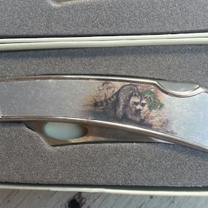 May include: A silver pocket knife with a raccoon design on the handle. The knife is closed and has a silver blade.
