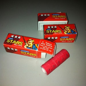 May include: Three red boxes with white stars and the text "Three Stars Roll Caps". The boxes contain rolls of red paper caps. The text on the box says "These Caps contain less than 20 grain per cap".