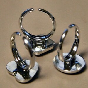 May include: Three silver-toned rings with open bands. The rings have a heart-shaped or square base. The rings are displayed on a neutral-colored surface. The rings are likely costume jewelry.