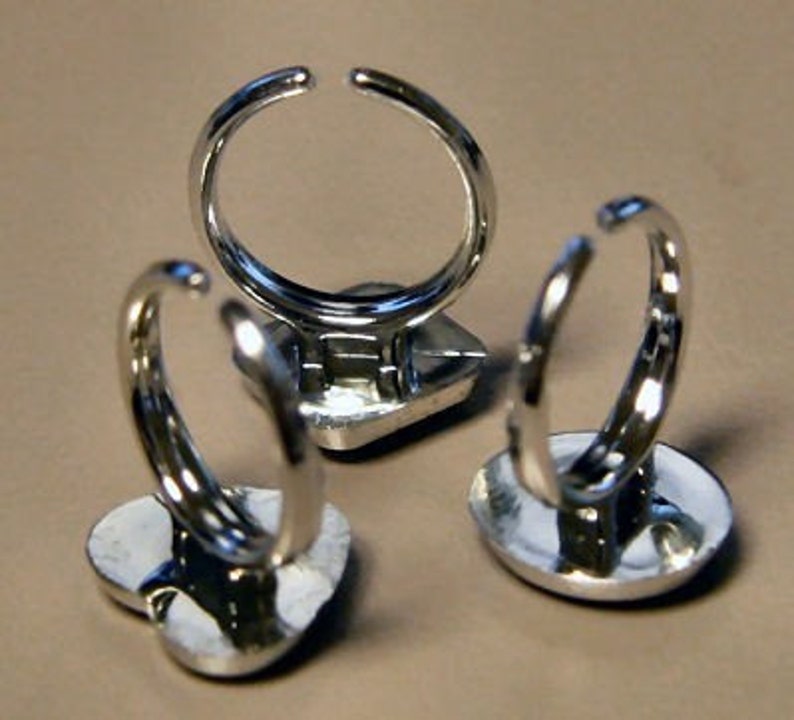 May include: Three silver-toned rings with open bands. The rings have a heart-shaped or square base. The rings are displayed on a neutral-colored surface. The rings are likely costume jewelry.