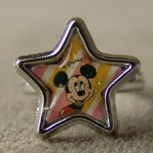May include: A silver-toned ring featuring a star-shaped face with a Mickey Mouse design. The face has a clear, protective layer over a colorful background with pink and yellow stripes. The ring is a children's accessory.