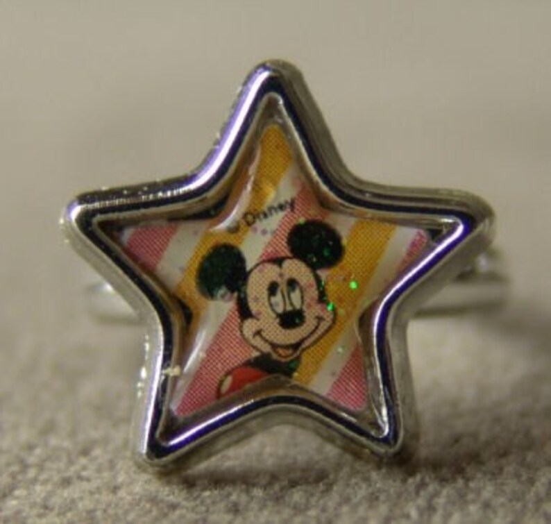 May include: A silver-toned ring featuring a star-shaped face with a Mickey Mouse design. The face has a clear, protective layer over a colorful background with pink and yellow stripes. The ring is a children's accessory.