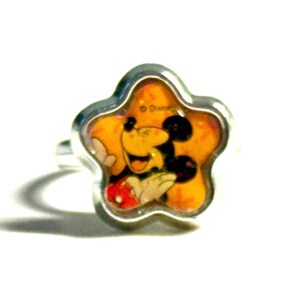 May include: A silver-toned ring featuring a star-shaped face with a smiling Mickey Mouse design. The ring has a yellow background with Mickey Mouse in his classic outfit. The ring is set against a white background.