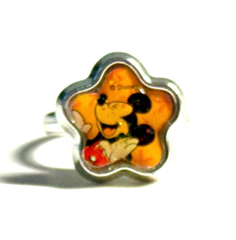 May include: A silver-toned ring featuring a star-shaped face with a smiling Mickey Mouse design. The ring has a yellow background with Mickey Mouse in his classic outfit. The ring is set against a white background.