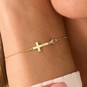 May include: A gold bracelet with a sideways cross pendant engraved with the name "Clare". A small, red gemstone is set near the clasp. The delicate bracelet is worn on the wrist. The cross is a religious symbol.