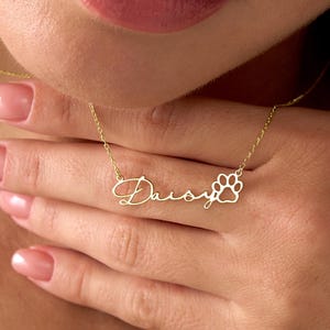 Pu&ograve; includere: Una delicata collana d'oro con il nome "Daisy" in corsivo, seguita da un ciondolo a forma di zampa. La collana &egrave; indossata intorno al collo. La catena e il ciondolo sono di colore oro.