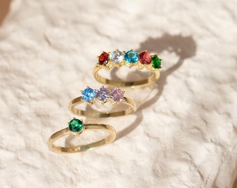 May include: Three gold rings with colorful gemstones. The top ring has five gemstones: red, white, blue, red, and green. The middle ring has three gemstones: blue, purple, and pink. The bottom ring has one green gemstone.
