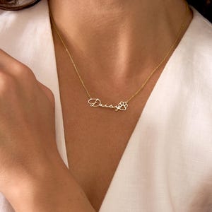 Pu&ograve; includere: Una collana color oro con il nome "Daisy" in corsivo, seguito da un ciondolo a forma di zampa. La collana &egrave; indossata intorno al collo, su un top bianco.