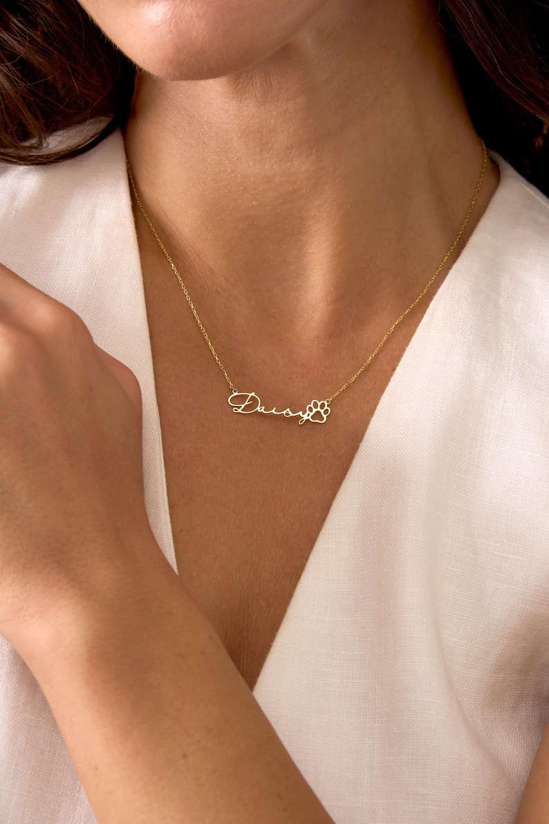 Pu&ograve; includere: Una collana color oro con il nome "Daisy" in corsivo, seguito da un ciondolo a forma di zampa. La collana &egrave; indossata intorno al collo, su un top bianco.