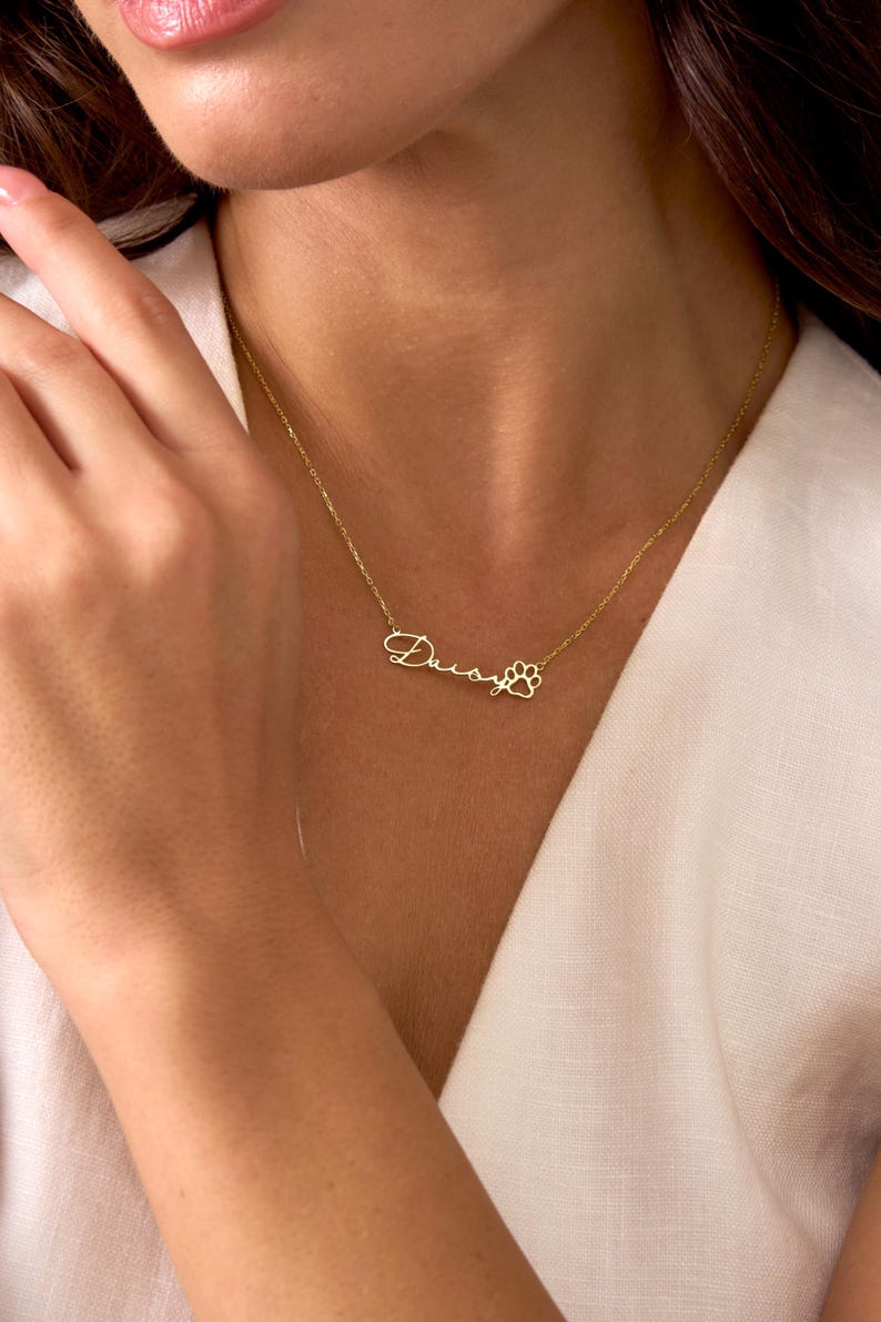Pu&ograve; includere: Una collana color oro con il nome "Daisy" in corsivo e un ciondolo a forma di zampa. La collana &egrave; indossata intorno al collo e poggia su un top bianco.