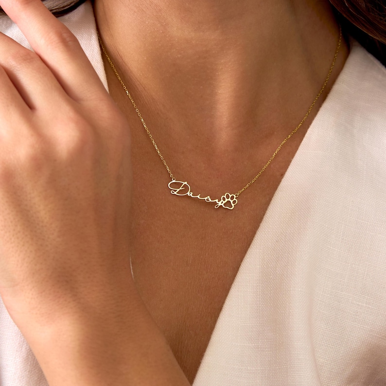 Pu&ograve; includere: Una delicata collana dorata con il nome "Daisy" in corsivo, seguito da un ciondolo a forma di zampa. La collana &egrave; mostrata su uno sfondo bianco, evidenziando il design e il colore del gioiello.