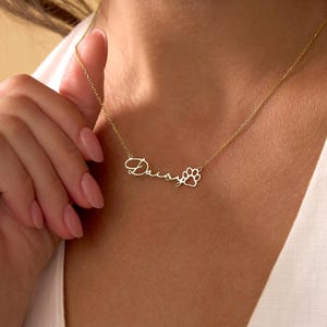 Pu&ograve; includere: Una collana color oro con il nome "Daisy" in corsivo e un ciondolo a forma di zampa. La collana &egrave; su una catena sottile ed &egrave; tenuta da una persona.