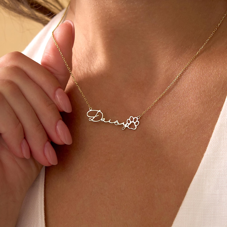 Pu&ograve; includere: Una collana color oro con il nome "Daisy" in corsivo e un ciondolo a forma di zampa. La collana &egrave; su una catena sottile ed &egrave; tenuta da una persona.