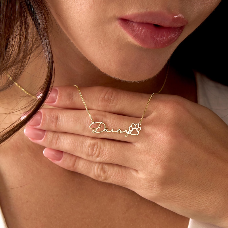 Pu&ograve; includere: Una collana color oro con il nome "Daisy" in corsivo e un ciondolo a forma di zampa. La collana &egrave; delicata e sembra essere in metallo. La catena &egrave; sottile e il ciondolo &egrave; centrato.