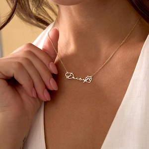 Pu&ograve; includere: Una collana color oro con il nome "Daisy" in corsivo e un ciondolo a forma di zampa. La collana &egrave; indossata intorno al collo. Lo sfondo &egrave; una persona che indossa un top bianco.