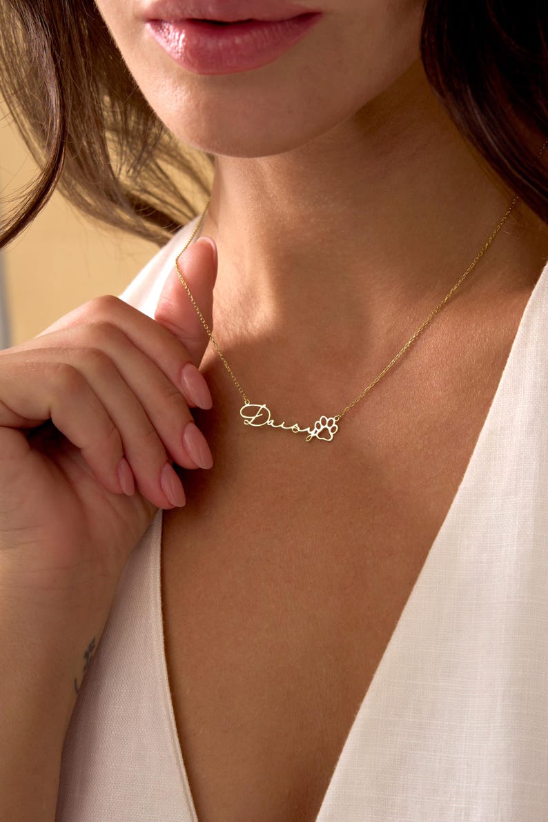 Pu&ograve; includere: Una collana color oro con il nome "Daisy" in corsivo e un ciondolo a forma di zampa. La collana &egrave; indossata intorno al collo. Lo sfondo &egrave; una persona che indossa un top bianco.