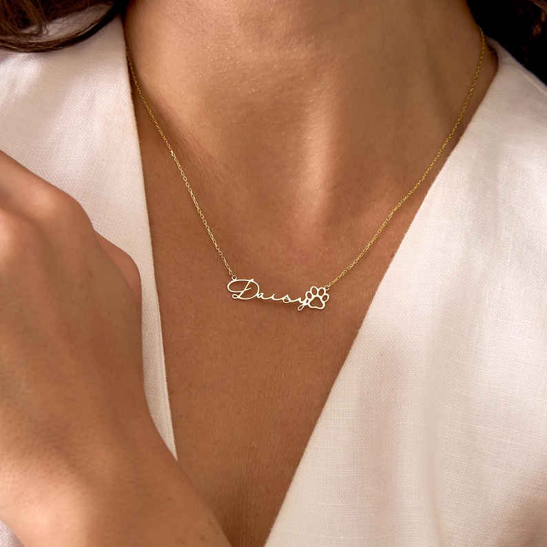 Pu&ograve; includere: Una collana color oro con il nome "Daisy" in corsivo, seguito da un ciondolo a forma di zampa. La collana &egrave; indossata intorno al collo e la catena &egrave; delicata.