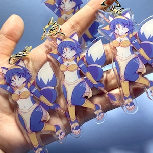May include: Clear acrylic keychains featuring a cartoon fox character with blue fur, white accents, and gold-colored armor. The fox has a determined expression and is posed in a dynamic stance. Gold star and other charms are also visible.