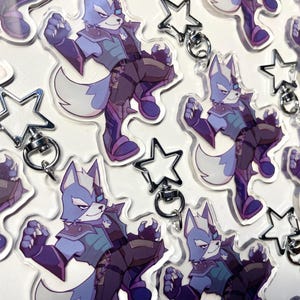 Puede incluir: Llaveros acrílicos transparentes con un personaje de lobo azul y morado en una pose dinámica. Cada llavero tiene un dije plateado en forma de estrella adjunto. El personaje lleva un atuendo oscuro con detalles de armadura.