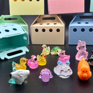 Op de afbeelding: Een verzameling kleurrijke, miniatuur dierenfiguren, waaronder een eend, schildpad, flamingo, eenhoorn, dolfijn en octopus. De figuren worden getoond voor kleine, pastelkleurige dozen.