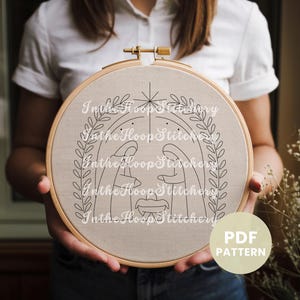 May include: A black and white embroidery pattern of the nativity scene with a star above the manger. The pattern is on a beige fabric hoop and the text "In the Hoop Stitchery" is repeated around the pattern.