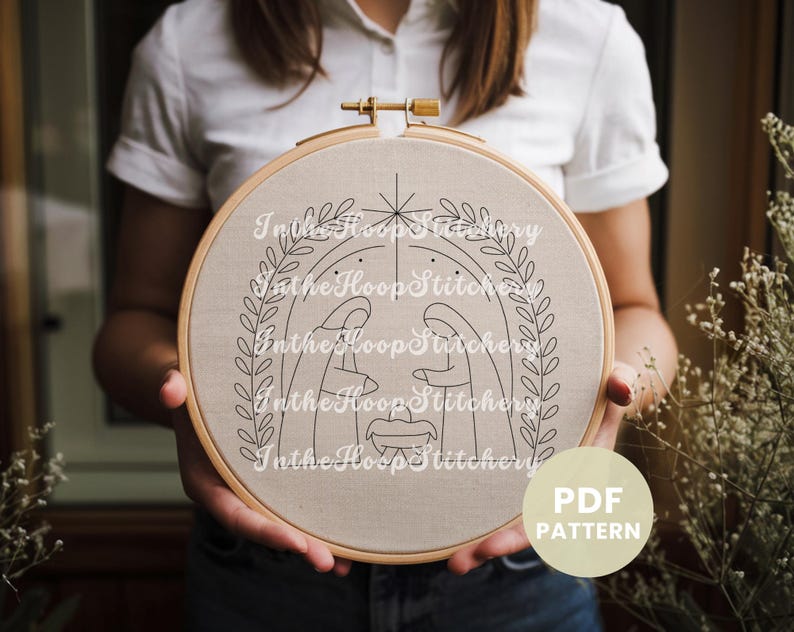 May include: A black and white embroidery pattern of the nativity scene with a star above the manger. The pattern is on a beige fabric hoop and the text "In the Hoop Stitchery" is repeated around the pattern.