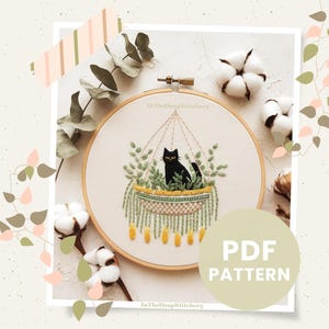 May include: A black cat sitting in a hanging planter with green plants and yellow tassels. The planter is in a wooden embroidery hoop. The text "PDF PATTERN" is on a beige circle in the lower right corner of the image. The text "In The Hoop Stitchery" is on the embroidery hoop.