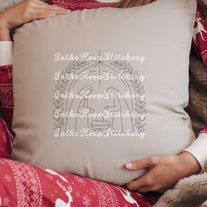 May include: A beige pillow with a black line drawing of the nativity scene inside a wreath. The text "In the Hoop Stitchery" is repeated around the image.