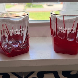 Può includere: Due bicchieri trasparenti con il logo bianco Maker's Mark stampato sul davanti. Ogni bicchiere è rivestito da una custodia in silicone rosso che imita l'aspetto della cera che cola, estendendosi dalla base lungo i lati del bicchiere.