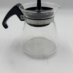 Op de afbeelding: Een glazen koffiepot met een zwarte handgreep en deksel. De pot heeft een afgeronde basis en een smalle hals. De handgreep zit aan de linkerkant en het deksel heeft een zwarte knop. Ontworpen om koffie te zetten.