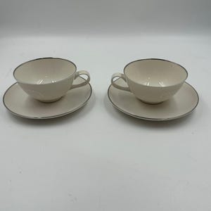 May include: Two cream-colored teacups and saucers. Each cup has a rounded shape with a handle and sits on a matching saucer. A thin silver-colored trim outlines the rims of the cups and saucers. The set is displayed on a white surface.