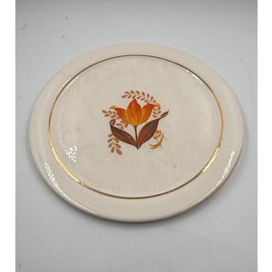 Könnte beinhalten: Cremefarbene Keramikplatte mit Goldrand und floralem Design. Das zentrale Design zeigt eine orangefarbene Tulpe mit braunen Blättern und Akzenten. Ein dekoratives Stück zum Servieren oder Ausstellen.