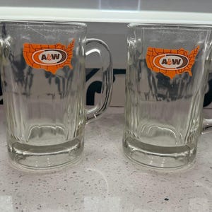 May include: Two clear glass mugs with sturdy handles and vertical ribbed detailing at the base. Each mug features an orange graphic of the United States map with the white and brown A&W logo printed in the center of the map design on the front.