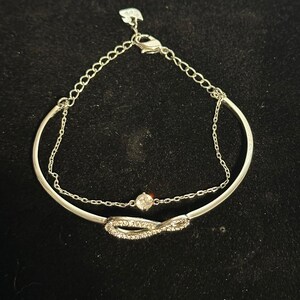 Puede incluir: Pulsera plateada con dos capas. La capa superior es una cadena fina, y la capa inferior presenta un diseño de símbolo de infinito incrustado con pequeñas piedras transparentes. Una pequeña piedra transparente y redonda está centrada en la pulsera.