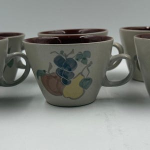 Può includere: Set di sei tazze da tè in ceramica con esterno beige e interno marrone scuro. Ogni tazza presenta un'illustrazione colorata di frutta, tra cui uva, pere e mele. Le tazze hanno un manico arrotondato e una forma leggermente svasata.