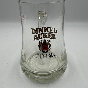 May include: Clear glass beer mug with a handle. The mug features the words "DINKEL ACKER" in black and red lettering, along with a logo and the text "CD-Pils".