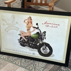 May include: Framed Harley-Davidson print featuring a pin-up girl on a green motorcycle. The print includes the text "American Bombshell" and "A salute from the home front to those who defend freedom."