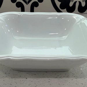 May include: A square white ceramic serving bowl with a smooth, glossy finish. The bowl features a wide, slightly raised rim with a subtle, molded decorative border detail along the edges, creating a clean and uniform appearance for the dish.
