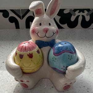 May include: A white ceramic Easter bunny figurine holding two egg-shaped salt and pepper shakers. The bunny has pink accents on its ears and cheeks, and a blue bow tie. The egg shakers are decorated with floral patterns.