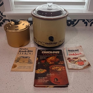 May include: A beige Rival Crock-Pot slow cooker with a glass lid, a gold-colored metal Crock-Ette, and three instructional booklets titled Crock-Ette Cooking, Crock-Pot Cooking, and Crock-Pot cookbook, all displayed on a white countertop.