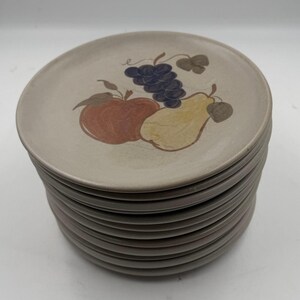 May include: A stack of beige ceramic plates with a fruit design. Each plate features a painted apple, pear, and grapes in shades of red, yellow, and purple. The plates have a brown rim.