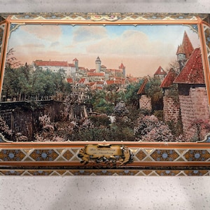May include: A decorative rectangular box with a detailed landscape of a European town, featuring red-roofed buildings and a castle. The box has an ornate border with gold, blue, and brown accents. A small plaque with text is visible.