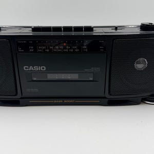 Könnte beinhalten: Ein schwarzer, tragbarer Casio Kassettenrekorder mit Radio, zwei seitlichen Lautsprechern, einem zentralen Kassettendeck und einem analogen Radiotuner. Das Gerät verfügt über einen Tragegriff, Bedientasten und eine Bass-Boost-Beschriftung.
