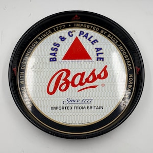 Puede incluir: Una bandeja redonda de metal negro para servir Bass Pale Ale. La bandeja presenta el logotipo de Bass en rojo, un triángulo rojo y el texto "Since 1777" e "Imported from Britain".