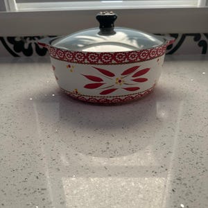 May include: White and red floral patterned cooking pot with a glass lid and black handle. The pot has a decorative red border and red and yellow floral designs. The pot is sitting on a white countertop.