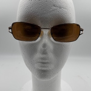 May include: Brown rectangular sunglasses with a thin, silver-colored metal frame. The lenses are a warm brown color. The sunglasses are displayed on a white foam head.