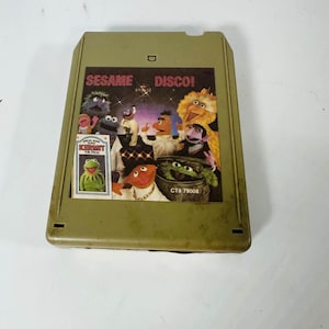 Op de afbeelding: Een olijfgroene 8-track tape met Sesamstraat-personages. Op het label staat "Sesame Disco!" met afbeeldingen van Big Bird, Kermit de Kikker en andere personages. De tape heeft een kleine sticker van Kermit de Kikker.