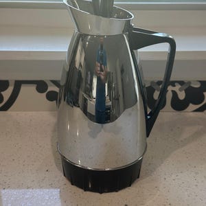 May include: A vintage stainless steel coffee carafe with a black base and handle. The carafe has a reflective surface and a lid with a black knob. The carafe is sitting on a white countertop.