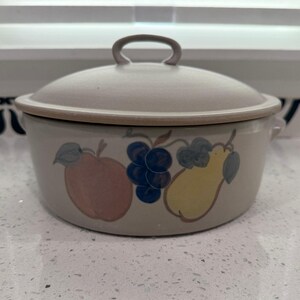 Può includere: Una pirofila in ceramica beige con coperchio. Il piatto è decorato con frutta dipinta tra cui una mela, uva e una pera. Il coperchio ha una maniglia. Il piatto ha due maniglie laterali.
