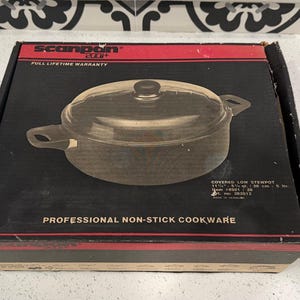 Könnte beinhalten: Eine schwarze Box mit roter Umrandung zeigt einen bedeckten niedrigen Kochtopf. Der Topf hat einen Deckel aus klarem Glas und zwei Griffe. Auf der Box steht "SCANPAN" und "PROFESSIONAL NON-STICK COOKWARE".